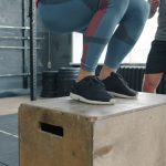 Low shot of sportswoman jumping onto wooden box during sports training with coach while man standing and watching. People, lifestyle and fitness concept.