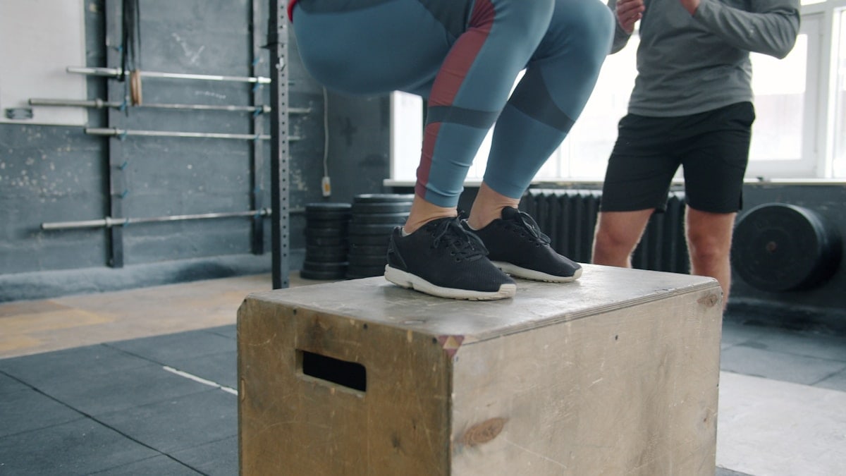 Low shot of sportswoman jumping onto wooden box during sports training with coach while man standing and watching. People, lifestyle and fitness concept.