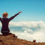 A woman enjoys a scenic view atop a cliff in Kula, Hawaii, surrounded by clouds and blue sky.